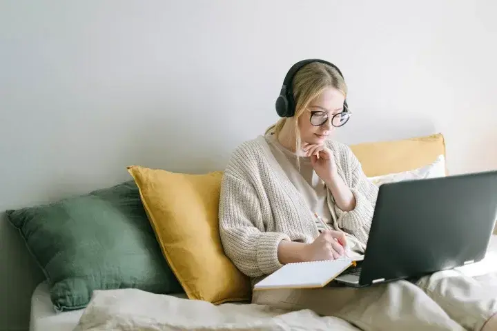 A women sits on a couch using a laptop while making notes.